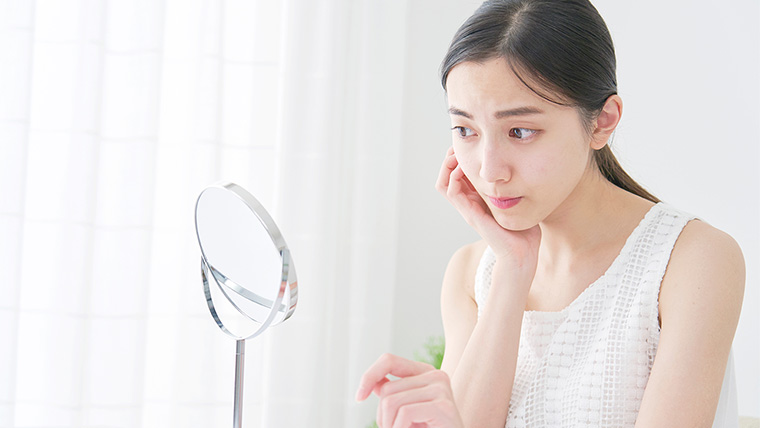 鏡の前で頬に手を添え、自分の肌の状態を真剣な表情で確認する女性。